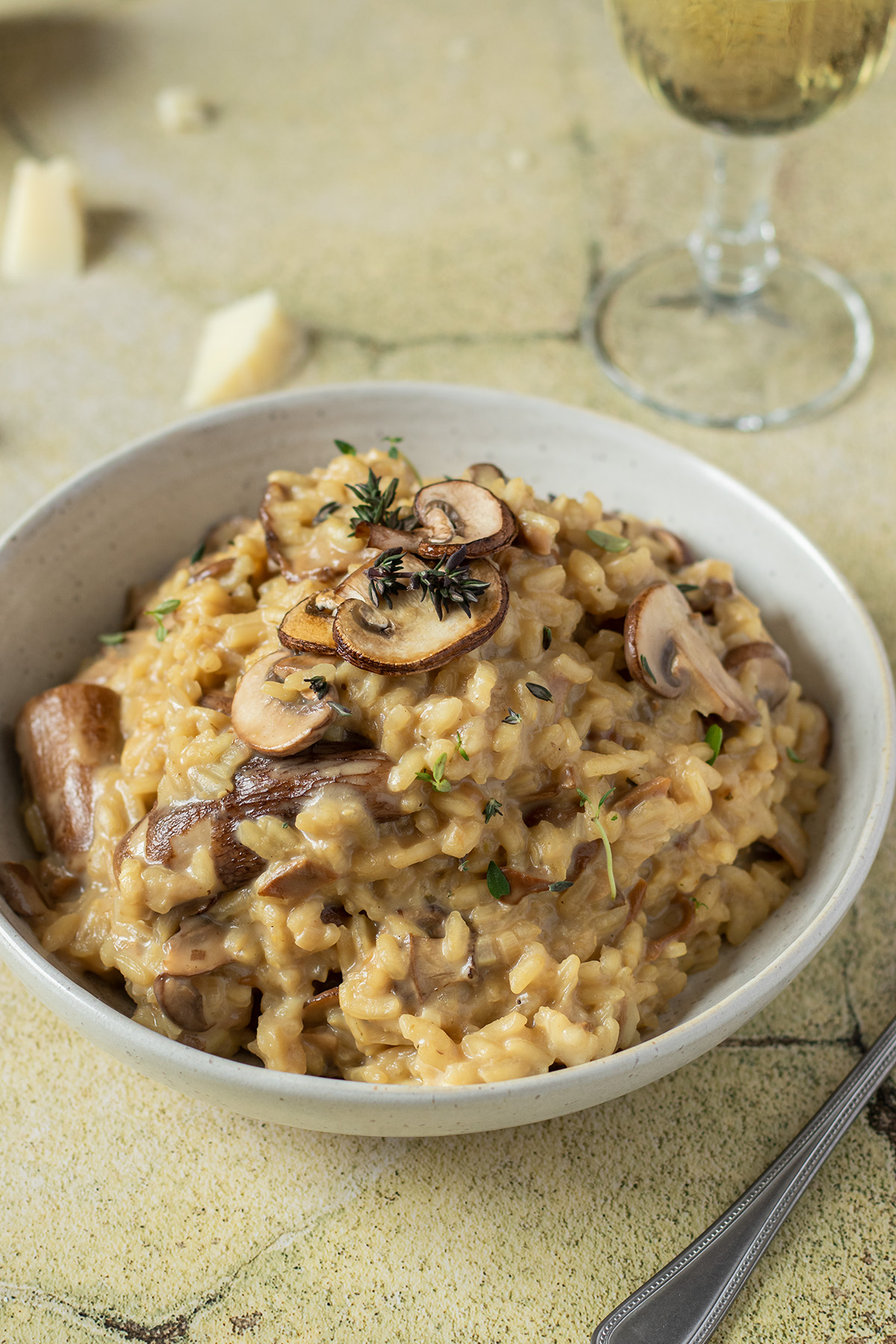 Teller mit cremigen Steinpilzrisotto auf grünen Fließen, Parmesan und ein Weinglas im Hintergrund.