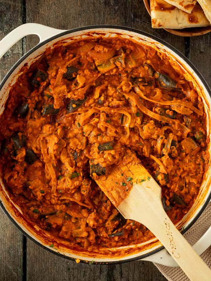 Auberginen Curry mit Linsen und Zwiebeln in einer cremigen Tomatensauce in einer gro&szlig;en Pfanne auf einem rustikalen Holzuntergrund, im Bildrand Naan.