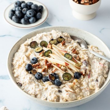 Bircher Müsli mit frischem Obst und Nüssen in einer Schüssel, frische Blaubeeren und Nüsse im Hintergrund.