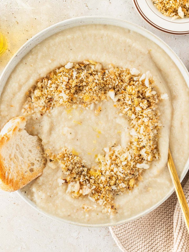 Teller mit Blumenkohlsuppe und einem Brotkr&uuml;mel-Topping in dem ein St&uuml;ck Brot getunkt wird, Deko im Bildrand.