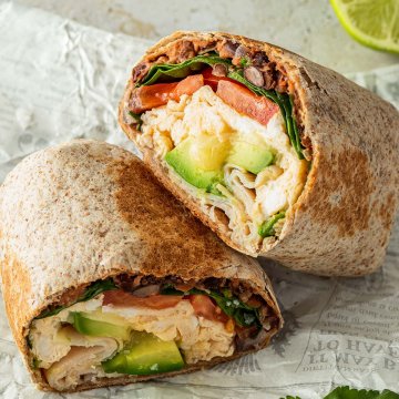 Halbierter Breakfast Burrito mit Rührei, Bohnen, Avocado, Spinat und Tomaten Füllung auf einem Stück Zeitungspapier. Deko im Hintergrund.