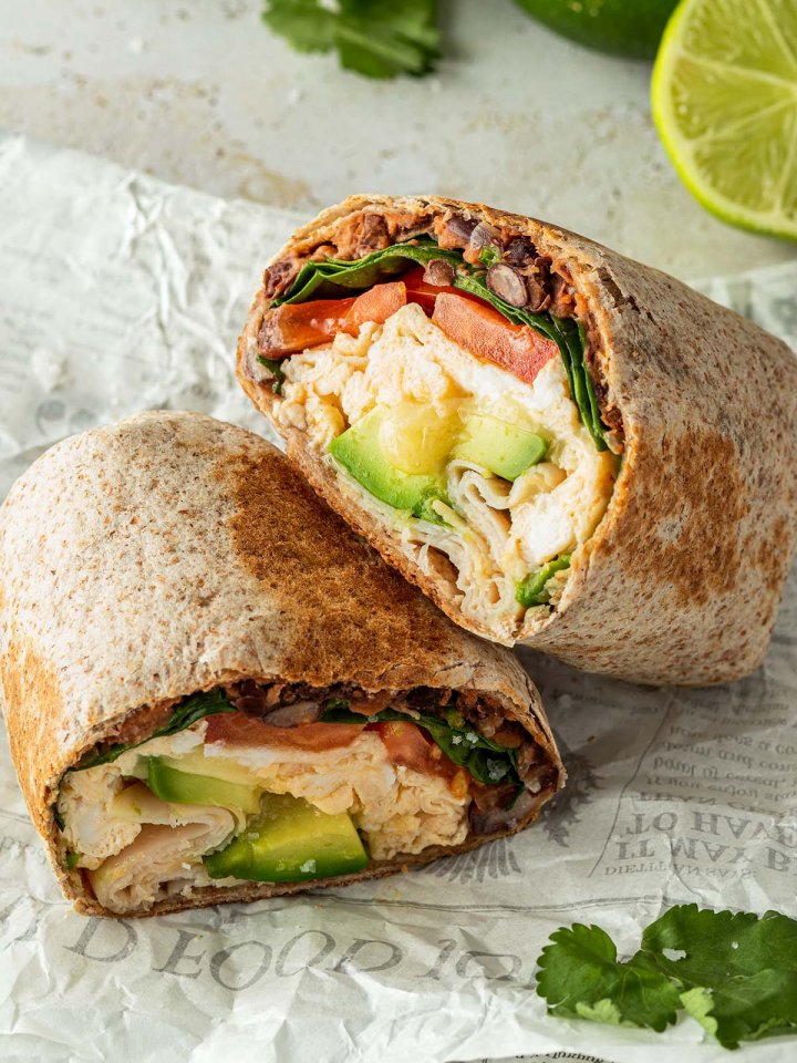 Halbierter Breakfast Burrito mit R&uuml;hrei, Bohnen, Avocado, Spinat und Tomaten F&uuml;llung auf einem St&uuml;ck Zeitungspapier. Deko im Hintergrund.