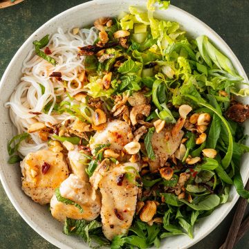 Eine Schüssel Bun Tron, ein vietnamesischer Reisnudelsalat mit frischen Kräutern, Salat, Hähnchen und knusprigen Erdnüssen, auf einem dunkelgrünen Untergrund.