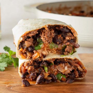 Ein in der Mitte halbierter Burrito gef&uuml;llt mit einer Hackfleisch und Bohnen Pfanne auf einem Holzbrett, im Hintergrund eine Pfanne mit Chili con Carne F&uuml;llung und frische Kr&auml;uter.