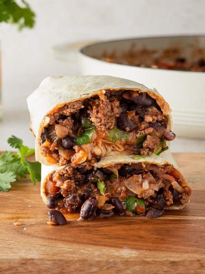 Ein in der Mitte halbierter Burrito gef&uuml;llt mit einer Hackfleisch und Bohnen Pfanne auf einem Holzbrett, im Hintergrund eine Pfanne mit Chili con Carne F&uuml;llung und frische Kr&auml;uter.