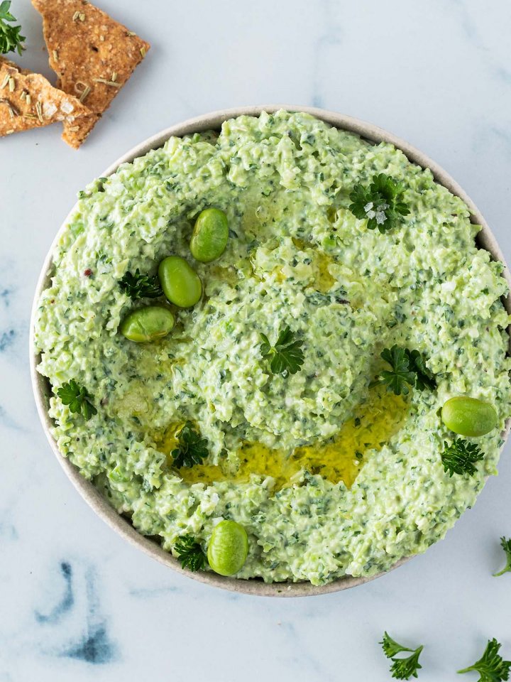 Vogelperspektive auf einen Edamame Dip, mit Cracker und Petersilie im Bildrand.