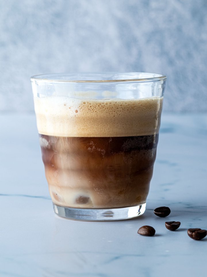 Kalt und schaumig gesch&uuml;ttelter Espresso in einem Glas vor blau-grauem Hintergrund.