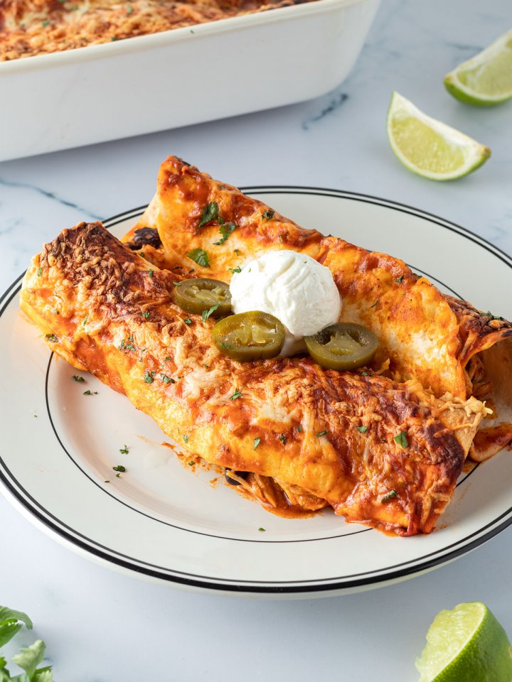 Zwei mit Käse überbackene Enchiladas auf einem Teller mit Sourcream und Jalapeños serviert, Deko und eine Auflaufform mit weiteren Enchiladas im Bildrand.