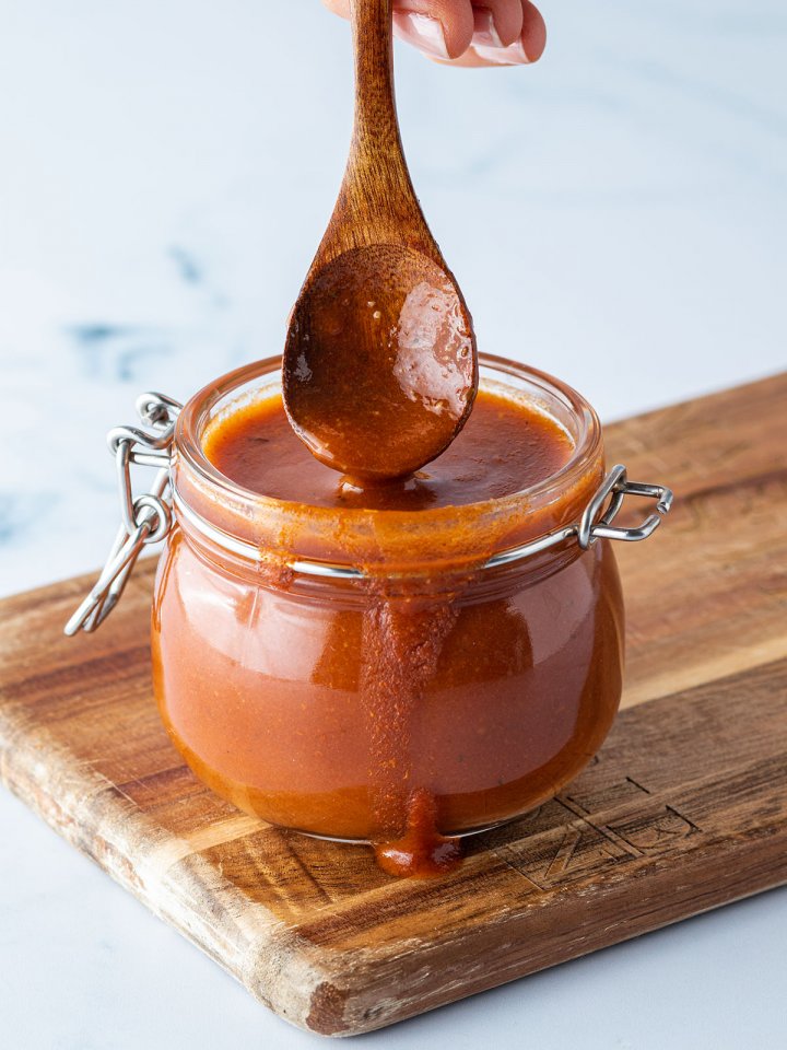 L&ouml;ffel wird in ein Glas mit einer dickfl&uuml;ssigen Enchilada-Sauce getaucht, das auf einem Holzbrett steht.