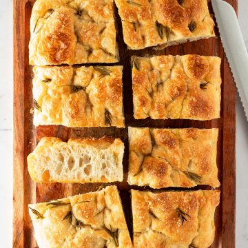 In rechteckige St&uuml;cke geschnittene Focaccia mit Rosmarin und grobem Salz auf einem Holzbrett, daneben ein Brotmesser.