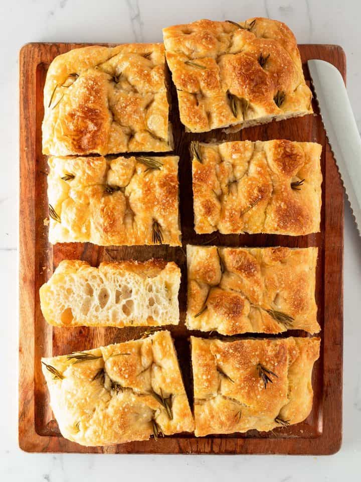 In rechteckige Stücke geschnittene Focaccia mit Rosmarin und grobem Salz auf einem Holzbrett, daneben ein Brotmesser.
