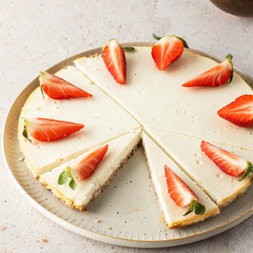 Ein Käsekuchen serviert auf einem Teller mit frischen, geviertelten Erdbeeren, eine Schale Erdbeeren im Hintergrund.