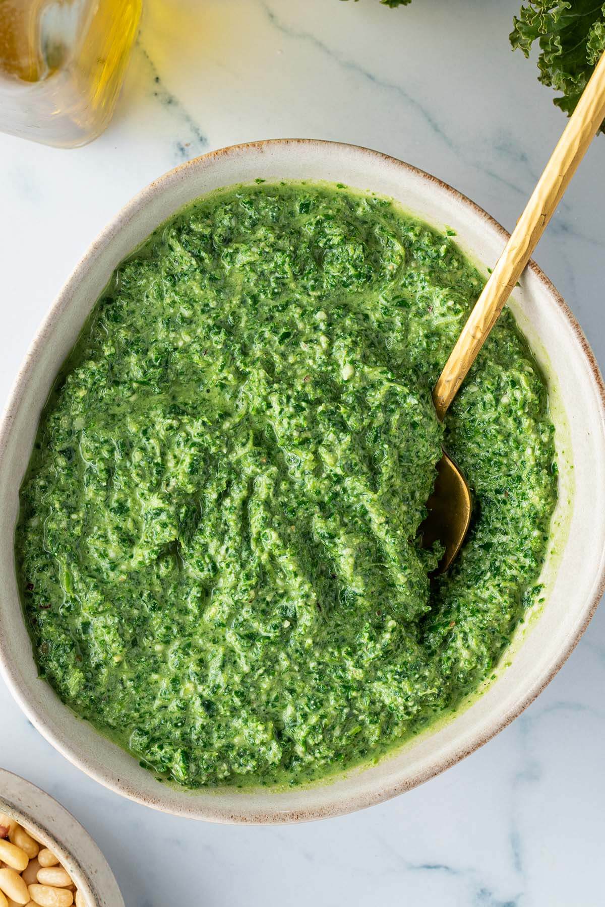 Schüssel mit einer Grünkohl Pesto auf einem Marmor-Untergrund, mit einer Flasche Öl und einer Schüssel Pinienkerne im Bildrand.