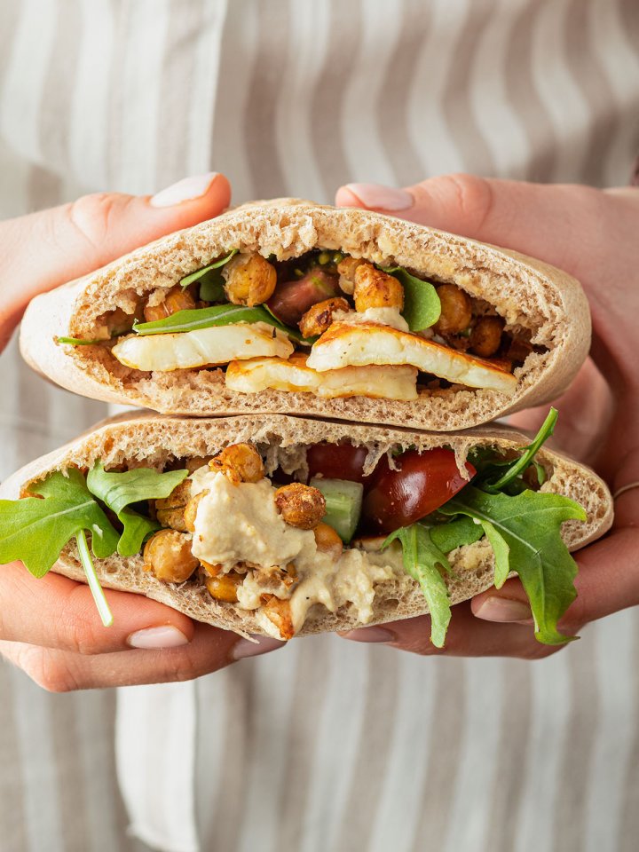 Eine Person mit Schürze hält zwei mit Halloumi, Kichererbsen und Hummus gefüllte Pita Taschen in der Hand.