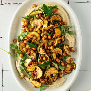 Hummus Platte mit ger&ouml;steten Kichererbsen, Zucchini und Rucola, Pita-Dreieche im Bildrand.