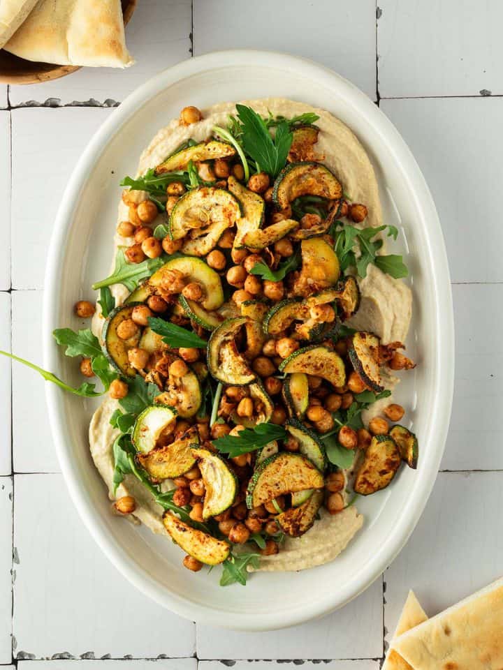 Hummus Platte mit ger&ouml;steten Kichererbsen, Zucchini und Rucola, Pita-Dreieche im Bildrand.