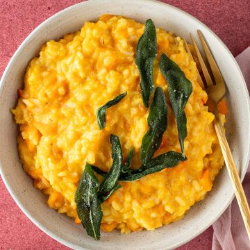 Cremige, oranges Kürbis Risotto mit knusprigen Salbei auf einem roten Untergrund, Deko im Bildrand.