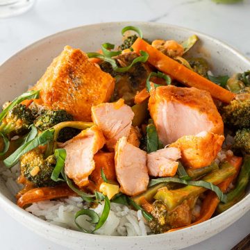Eine Schüssel mit Thai Lachs Curry mit saftigen Lachswürfeln, Karotten und Brokkoli serviert auf Reis mit frischem Basilikum. Im Hintergrund ein Glas Wasser, eine Limette und Esstäbchen im Bildrand.
