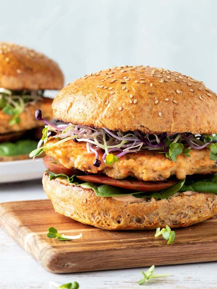 Ein saftiger Lachsburger mit einem goldbraun angebratenen Lachs Patty, frischen Sprossen, Tomaten und Salat auf einem Holzbrett, im Hintergrund ein zweiter Burger.