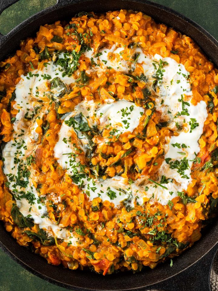 Nahaufnahme von einem Linsen Dal mit Spinat, da in einer Pfanne mit Joghurt und frischen Kr&auml;utern serviert wird.