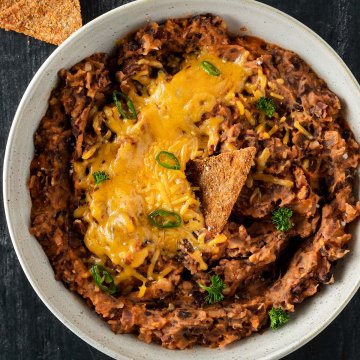 Mexikanisches Bohnenmus aus schwarzen Bohnen in einer Schüssel mit geschmolzenem Käse on Top und Chips dazu.