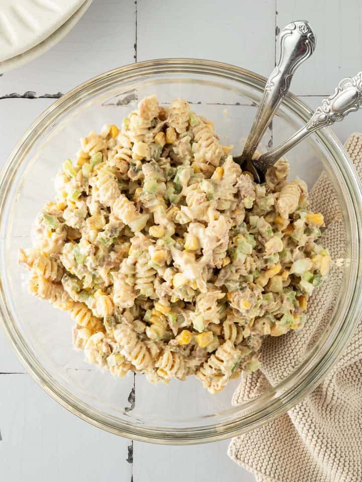 Ein cremiger Nudelsalat mit Thunfisch, Mais und Gemüse in einer großen Schüssel, Teller und ein Tuch im Bildrand.