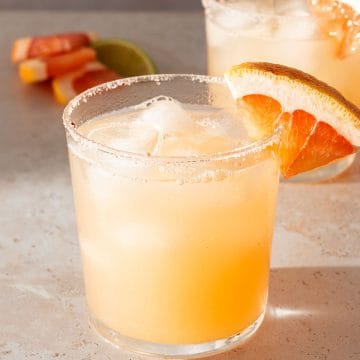 Paloma Cocktail in einem Glas mit Salzrand und einer Grapefruit-Scheibe, im Hintergrund ein zweites Glas.