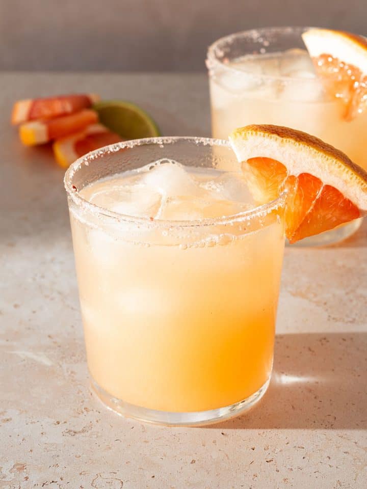 Paloma Cocktail in einem Glas mit Salzrand und einer Grapefruit-Scheibe, im Hintergrund ein zweites Glas.