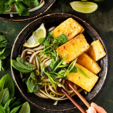 Zwei Sch&uuml;sseln vegetarischer Pho Suppe mit Nudeln, Kr&auml;utern und knusprigen Tofuscheiben auf einem dunkelgr&uuml;nen Untergrund und einer Hand, die zwei St&auml;bchen h&auml;lt.
