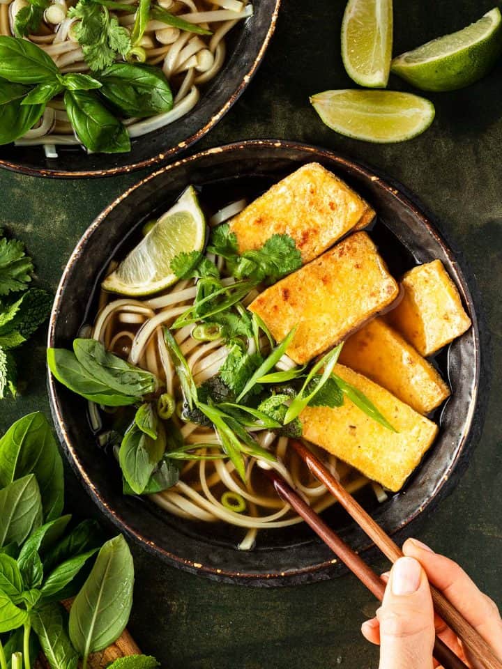 Zwei Sch&uuml;sseln vegetarischer Pho Suppe mit Nudeln, Kr&auml;utern und knusprigen Tofuscheiben auf einem dunkelgr&uuml;nen Untergrund und einer Hand, die zwei St&auml;bchen h&auml;lt.
