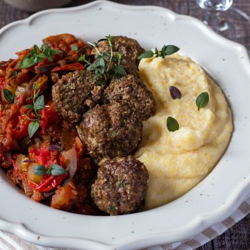 Cremige Polenta serviert mit Hackbällchen und Tomatengemüse in einem weißen Teller, Deko im Hintergrund.