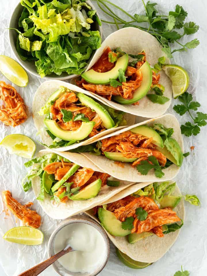 Fünf Tacos mit Pulled Chicken, Avocado und Salat auf einem weißen Untergrund mit frischen Kräutern, Salat und saurer Sahne.
