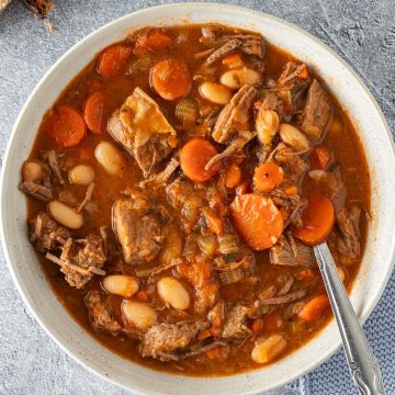Eine Sch&uuml;ssel Rindfleischeintopf mit Karotten und wei&szlig;en Bohnen auf einem grauen Untergrund, zwei Scheiben Brot im Bildrand.