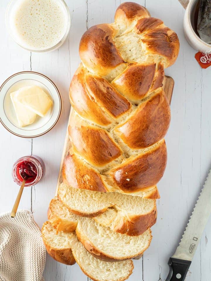 Ein weißer Holztisch mit einem angeschnittenen Hefezopf, außen herum Tassen, Butter, Marmelade und weitere Frühstücksutensilien.