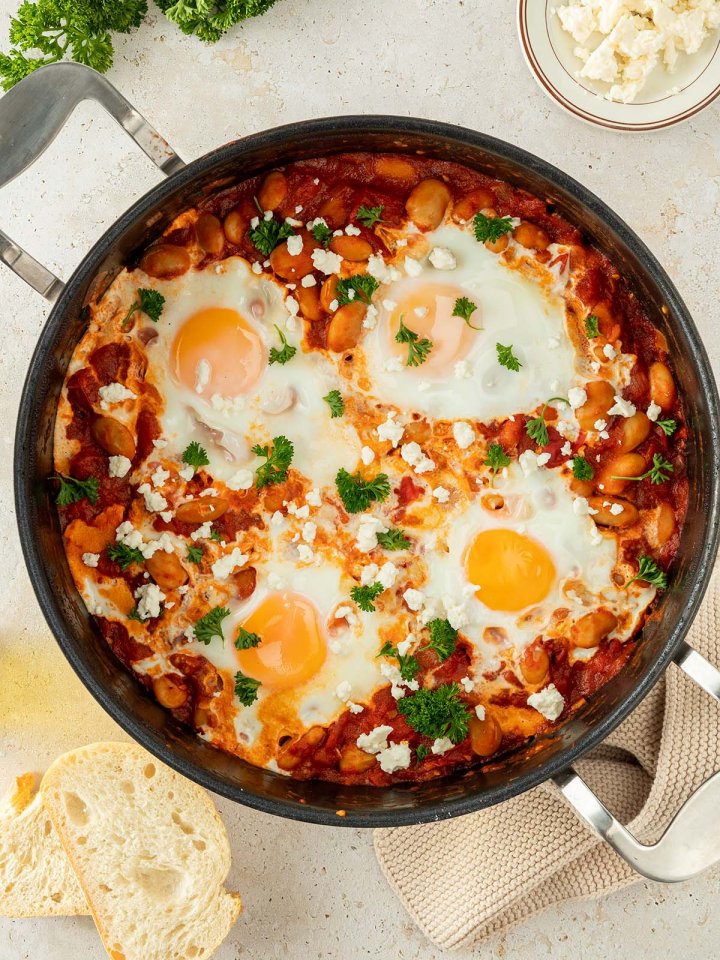 Vogelperspektive auf eine Pfanne mit Shakshuka, das mit frischer Petersilie und Kr&auml;utern serviert wird, Deko im Bildrand.