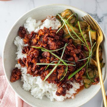 Bowl mit weißem Kokosreis, knusprig gebratenem Sticky Tempeh und würzigem Gurkensalat, garniert mit Frühlingszwiebeln und serviert mit einer Gabel.