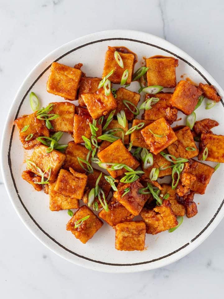 Knusprige Tofu-Stücke mit einer Sticky süß-sauer Sauce auf einem Teller mit Frühlingszwiebeln.