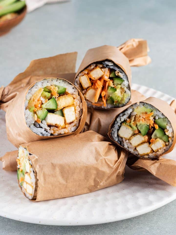Mehrere, halbierte und in Papier eingewickelte Sushi Burritos mit Tofu auf einem Teller.