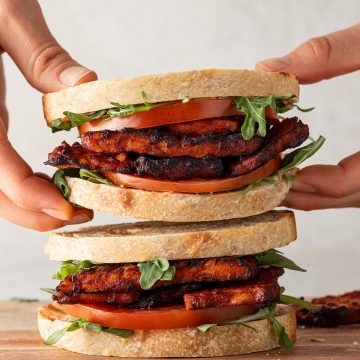Jemand nimmt eines von zwei übereinander gestapelten Tempeh Sandwiches weg.