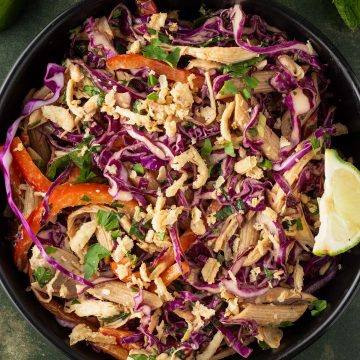 Thai Crunch Salat mit Erdnussdressing in einer schwarzen Schüssel auf dunkelgrünem Untergrund, Deko im Bildrand.