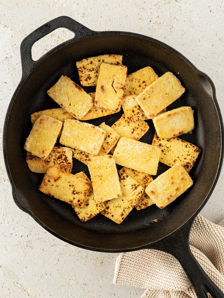 Knusprig gebratener Tofu in einer schwarzen Pfanne auf einem beigen Untergrund.