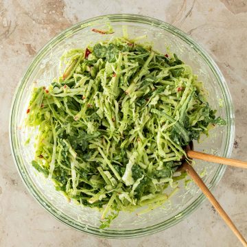 Eine Schüssel mit Weißkohlsalat mit Apfel, Rucola und grünem Dressing auf einem Steinuntergrund mit Salatbesteck.